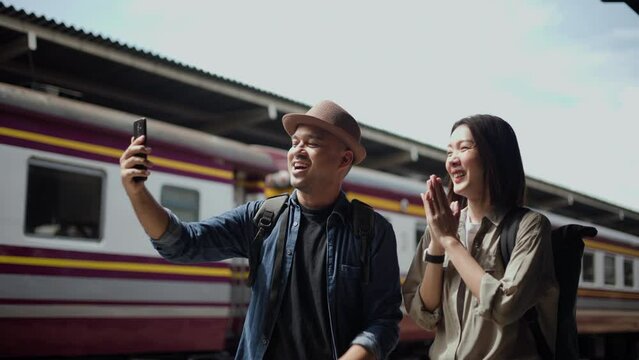 Couple asian traveller taking photo by smartphone while travel by train. Happy Man and woman standing near platform train station take a selfie. Planning tourist on vacation time holiday weekend.