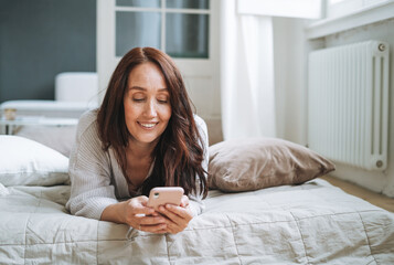 Young woman forty year with brunette long hair in cozy knitted cardigan using mobile phone in bed at home