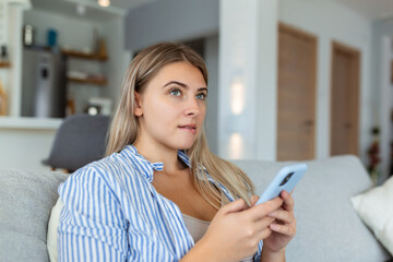 Image of nice young serious woman using mobile phone and thinking while sitting on sofa at living room