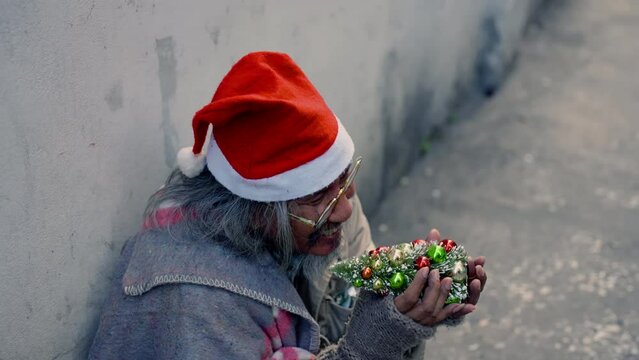 Asian homeless old man wearing christmas hat and holding small christmas tree sitting on wayside with hope, dirty alcoholism aged man with beard on street begging for money, no job.
