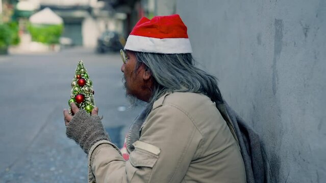 Asian homeless old man wearing christmas hat and holding small christmas tree sitting on wayside with hope, dirty alcoholism aged man with beard on street begging for money, no job.
