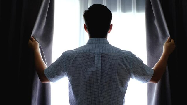 Man in shirt stands at window and opens curtains, back view.