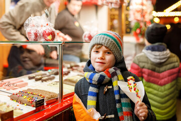 Little kid boy, cute child eating bananas covered with chocolate, marshmellows and colorful sprinkles near sweet stand with gingerbread and nuts. Happy boy on Christmas market in Germany.