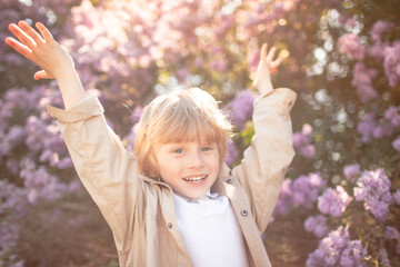 A little stylish boy dressed in a beige coat walks among flower trees in spring and the kidraaised  hand up