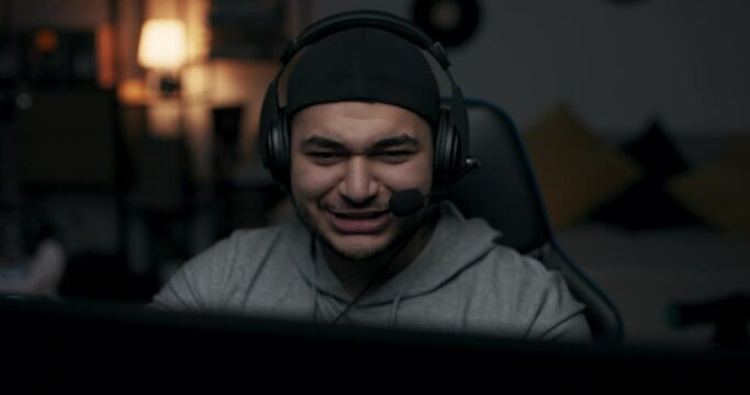 Bearded man in headphones is playing online game. He had been preparing for match for long time. Guy in gray T-shirt communicates with rivals. He wins, takes off headphones, claps hands and rejoices.