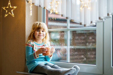 Little preschool girl holding cup with hot chocolate with marhsmallows. Happy child drinking sweet...