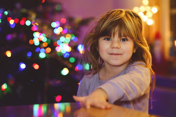 Litte toddler girl sitting in pajamas on Christmas morning or eve and with xmas tree and lights on background. Happy healthy child celebrate family traditional holiday, waiting for gifts.