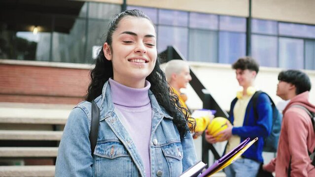 Portrait of young hispanic brunette female girl sanding out of the school. In the background three teenagers talking and laughing about the lessons learned in class. Education concept. High quality 4k