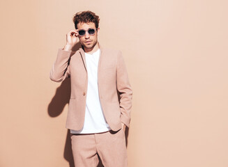 Portrait of handsome confident stylish hipster lambersexual model. Sexy modern man dressed in elegant suit. Fashion male with curly hairstyle posing in studio. Isolated on beige. In sunglasses