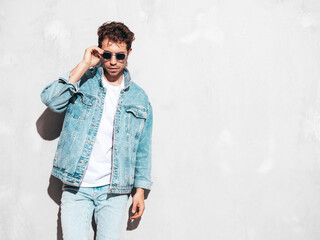 Portrait of handsome confident stylish hipster lambersexual model with curly hairstyle. Sexy man dressed in jeans jacket. Fashion male isolated on grey wall in studio. In sunglasses
