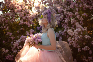 Portrait of a beautiful girl with a wreath on her head made of a purple flowers. A woman dressed pink skirt and blue sleeveless top walks outside against the background of flowering purple trees