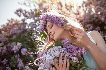 Fototapeta premium Portrait of a beautiful girl with a wreath on her head made of a purple flowers. A woman dressed pink skirt and blue sleeveless top walks outside against the background of flowering purple trees