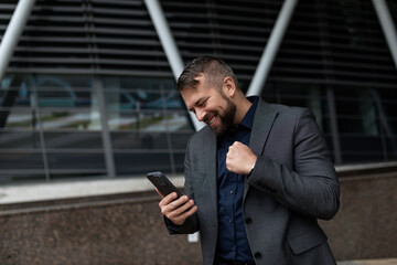 Naklejka na ściany i meble businessman in a classic suit on the background of an office building with a mobile phone in his hands energetic and emotional clenching his hand into a fist looking at the phone screen with a smile