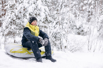 Portrait of teenage boy sitting on tubing winter snowy forest. Child walking resting relaxing...