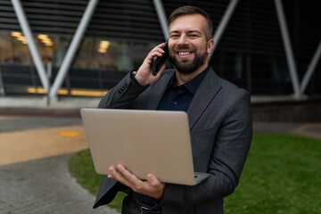 pretty adult man in a business suit with a laptop in his hands speaks on a mobile phone on the background of the office center, Concept of a successful realtor