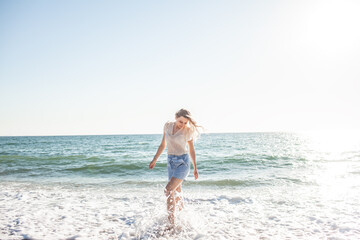 Fototapeta na wymiar A beautiful girl dressed in a blue shorts and a white light blouse runs on a deserted beach, enjoys freedom and loneliness and look at the biew of the ocean