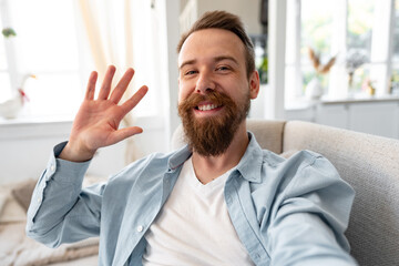 Bearded happy young man smiling and taking selfie photo while sitting on sofa at home.