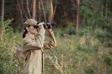 A woman park ranger in uniform looks through binoculars and monitoring the forest area in summer,...