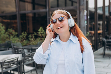 Lovely happy young woman in sunglasess listening to music through wireless earphones on urban background. Music lover enjoying music. Lifestyle concept. Girl walking on city street