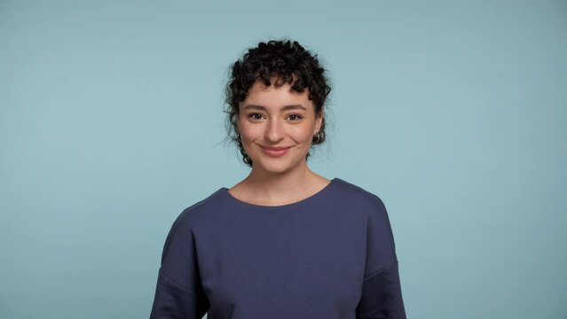 Close up smiling curly woman in blue sweater posing looks camera kisses courting flirting. Portrait positive female standing on isolated light blue background with copy space. People emotions concept
