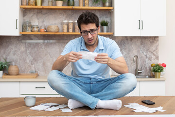 Stressed man sitting on the table and holding many bills in his hands in a spending, taxes and debt...