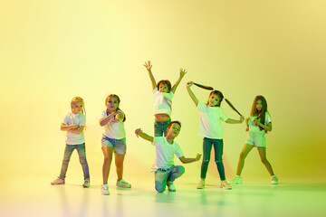 Stylish children, little girls and boys in casual style clothes dancing hip-hop isolated on green background in yellow neon light.