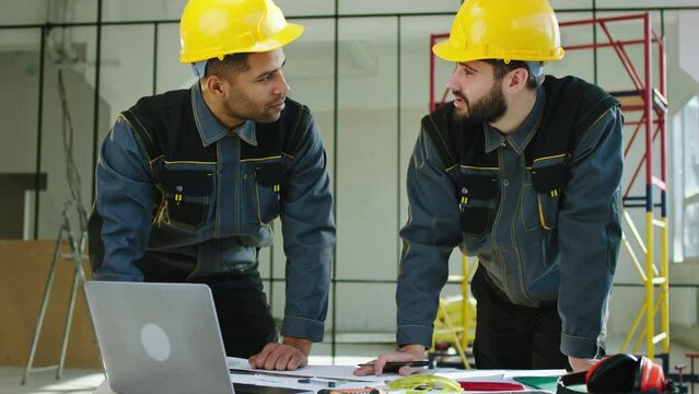 At construction site two young charismatic multiracial men analysing the plan of work using the laptop and plan of construction to analysing the construction building wearing safety equipment and