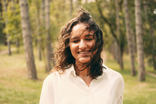 Portrait of young beautiful woman with curly hair standing in pine forest with clossed eyes in wind