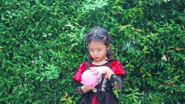 Small girl in Halloween Dress Playing With Pink Ball Against green Nature Background.