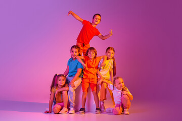 Dance group of happy, active little girls and boys in bright clothes in action isolated on pink background in neon. Concept of music, fashion, art, childhood