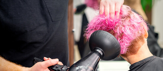 Drying short pink bob hairstyle of a young caucasian woman with a black hair dryer with the brush...
