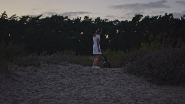 Wide view of young woman walking towards her dog on hill in sand dunes at dusk