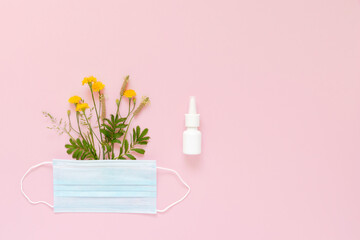 A set of wild flowers and herbs under a disposable protective mask and a mock-up of nasal spray on a light pink background. The concept of means of protection and treatment of seasonal allergies.