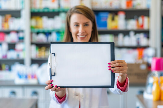 Smilin female pharmacist working in chemist shop or pharmacy. Female pharmacist in white uniform showing blank clipboard to write it on your personal message or advice.