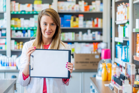 Smiling pharmacist standing in drug store with blank clipboard to write it on your personal message or advice. Young female pharmacist standing behind the counter with a clipboard