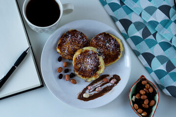Fototapeta Cheesecakes with chocolate paste, nuts, raisins, powdered sugar. A napkin, a mug of coffee, a notebook, a pen. Top view. obraz