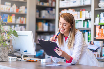 Medicine, pharmaceutics, health care and people concept - Smiling young pharmacist holding a clipboard and box of medications. Pharmacist writing on clipboard in pharmacy
