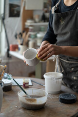 Female artisan enjoys process of handmade pottery craft standing at round wooden table with painting equipment. Woman in black apron creates white ceramic mug on blurred background in workshop closeup