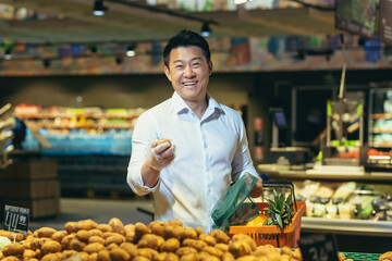 A young handsome Asian man, businessman is standing in a supermarket in the vegetable section, holding a basket of fruits and potatoes in his hands. He looks at the camera, smiles.