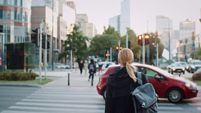 Young blond hair woman with backpack dressed in casual clothes standing by crossroad with lot of cars and pedestrians in city landscape. Female in busy street in evening, urban life, after work time