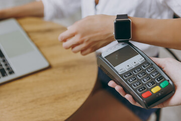Cropped shot young woman wear white shirt holding pay waiter smart watch modern wireless bank payment terminal sit at table in coffee shop cafe restaurant indoor work or study on laptop pc computer