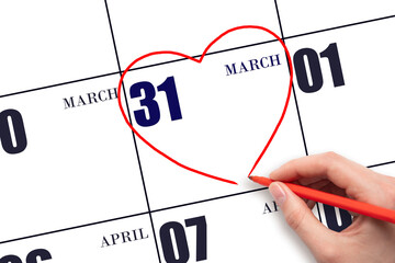 A woman's hand drawing a red heart shape on the calendar date of 31 March. Heart as a symbol of...