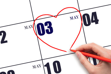 A woman's hand drawing a red heart shape on the calendar date of 3 May. Heart as a symbol of love.