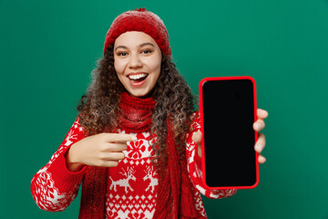 Merry young woman wearing red warm sweater hat posing hold in hand use show mobile cell phone blank screen workspace area isolated on plain dark green background. Happy New Year 2023 holiday concept.