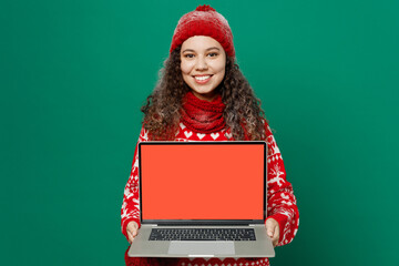 Merry young IT woman wear red warm knitted sweater hat posing hold use work on blank screen laptop pc computer isolated on plain dark green background. Happy New Year 2023 celebration holiday concept.