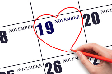 A woman's hand drawing a red heart shape on the calendar date of 19 November. Heart as a symbol of...
