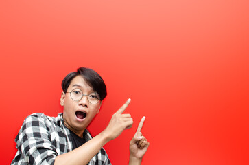 asian young man with glasses shocked happily pointing to copy space isolated over red background. billboard advertisment model concept.