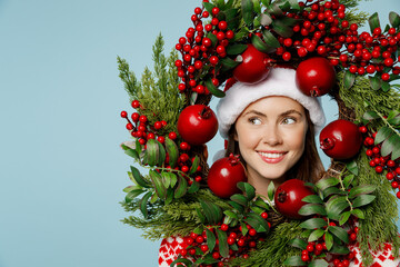 Young fun happy merry woman wear Santa hat posing look throught christmas wreath look aside on workspace area isolated on plain pastel light blue cyan background. Happy New Year 2023 holiday concept.