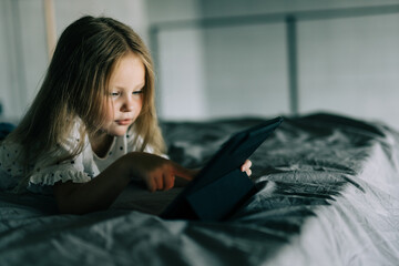 Fototapeta premium Little girl lying on bed look at screen of tablet at home