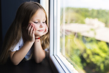 Beautiful little girl smiling and watching out the window. A child looks out the window. Portrait of cheerful kid lies at windowsill.